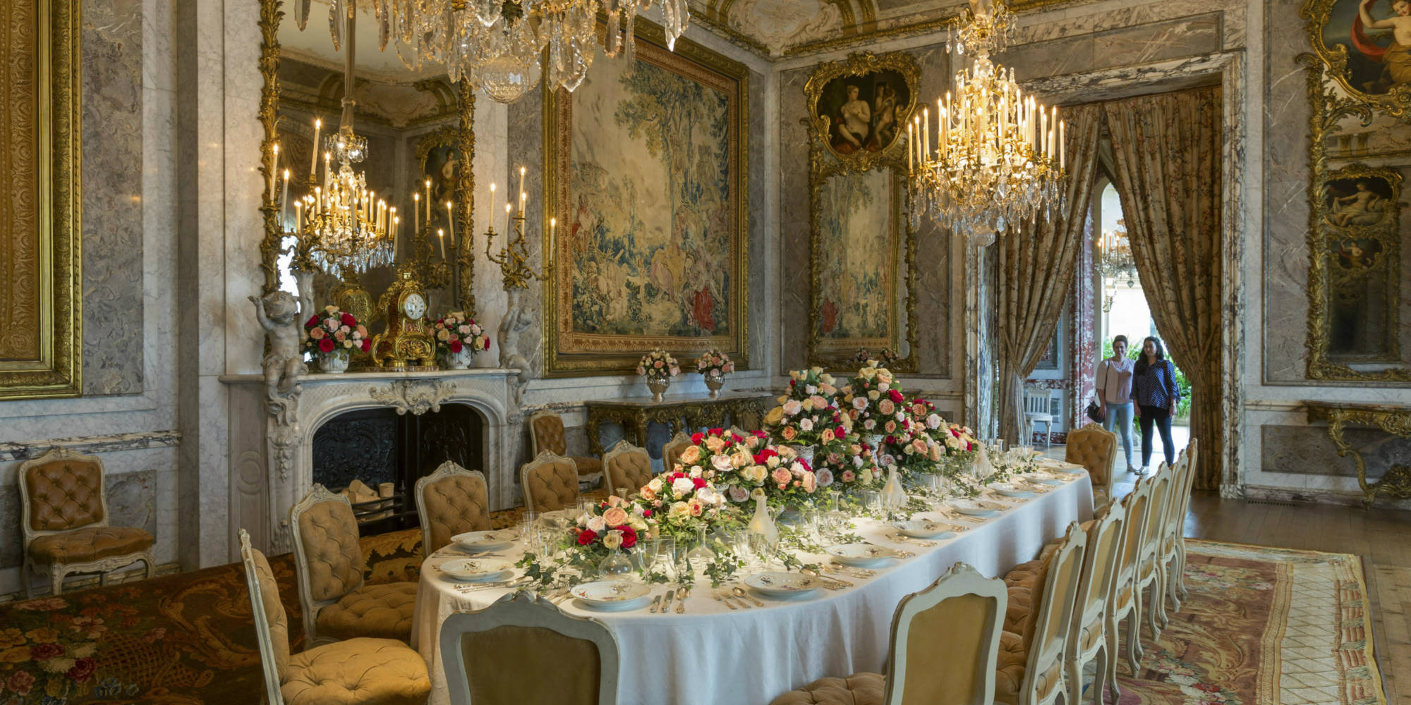 Long dining table covered in roses. Two visitors stand at the far end and admire the scene
